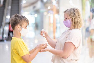 Woman wearing protective mask applies sanitizer for cleaning son's hands - in a shopping center or airport