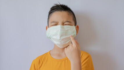 An Asian boy who is closing their eyes wears a COVID-19 virus mask and is dustproof. Raise his hand and point his mask. White background in the house