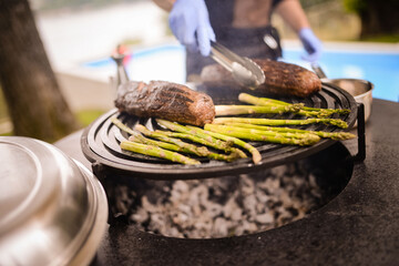 The chef grills steak and asparagus on the grill in the yard