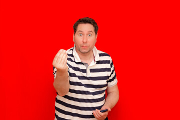 confusion man wearing horizontal striped t-shirt standing over red background doing money gesture with hands.