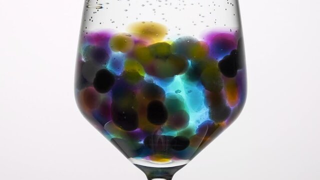 A timelapse of coloured orbeez, aka hydrogel, expanding in a glass of water against a white background. Slow zoom out from glass.