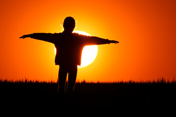 Fototapeta na wymiar A little girl stands on the meadow and watches the beautiful sunset with her hands outstretched. close up of the sun in the big orange sky.