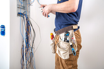 Electrician Builder at work, examines the cable connection in the electrical line in the fuselage of an industrial switchboard. Professional in overalls with an electrician's tool.