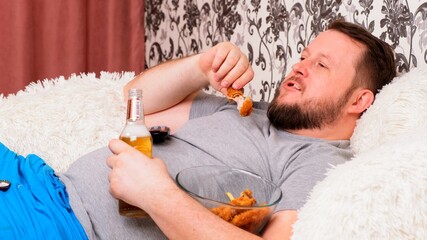 fat man drinks beer and eats unhealthy food chicken wings, bored in front of the TV outlook on the couch. The concept of malnutrition, quarantine at home, alcoholism.