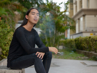 Young Asian man with long hair sitting and smoking at the park