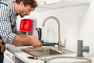 Side view of handsome repairman using pipe wrench while fixing kitchen faucet
