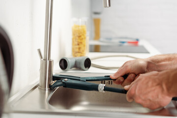 Cropped view of plumber fixing kitchen faucet with pipe wrench near pipes on worktop