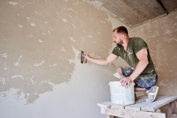 Young man at wooden stand is puttying the walls indoors. Guy with beard in t-shirt and jeans is...