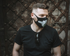 Handsome fashionable guy in a black t-shirt with designer protective black face mask with images of protest. Guy looks away. Trendy Fashion accessory during quarantine of coronavirus pandemic.