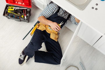 Top view of handyman in overalls and tool belt holding pliers while repairing sink in kitchen