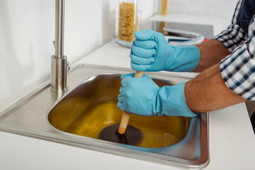 Cropped view of plumber in rubber gloves cleaning blockage of kitchen sink with plunger