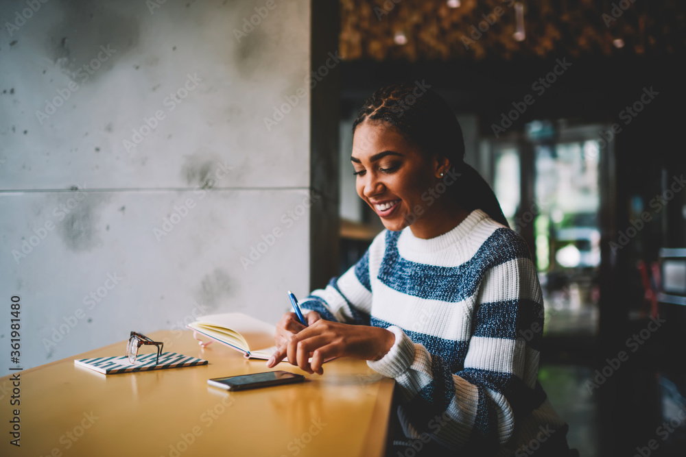 Poster cheerful afro american hipster girl writing down to do list in notepad while getting message on smar