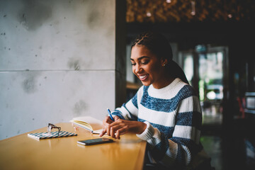 Cheerful afro american hipster girl writing down to do list in notepad while getting message on...