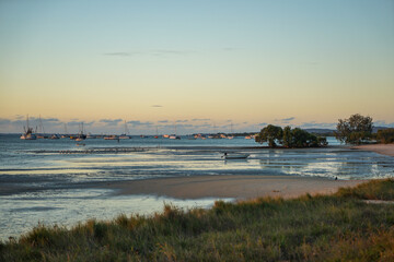 Obraz premium Twilight view of beach at low tide with grassy foreground, sand, trees, boats, and clouds on the horizon.