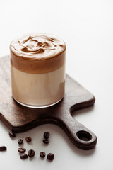 delicious Dalgona coffee in glass on wooden cutting board near coffee beans on white background
