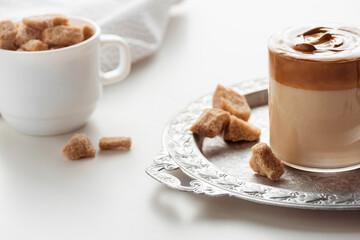 selective focus of delicious Dalgona coffee in glass near brown sugar on silver tray on white background