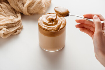 cropped view of woman holding spoon with foam near delicious Dalgona coffee in glass and cloth on white background