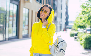 Stylish girl. A half-length photo of a smiley girl on the street, in yellow glasses , with smartwatch on her hand, silver backpack on her shoulder and hood on her head.