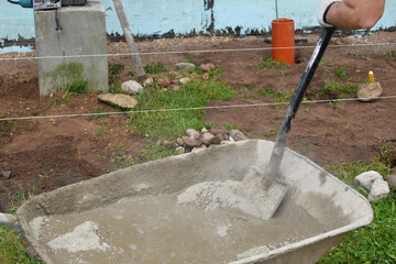 Worker hand mixing concrete mortar with shovel in old metal wheelbarrow on renovation site.
