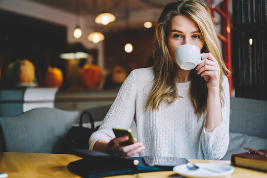 Portrait of attractive young woman with blonde hair drinking tasty aroma coffee in leisure time in cafeteria.Beautiful female enjoying beverage while looking at camera and holding telephone in hand