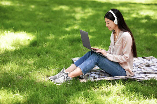 Smiling Asian Girl Relaxing With Laptop And Headphones On Lawn In Park