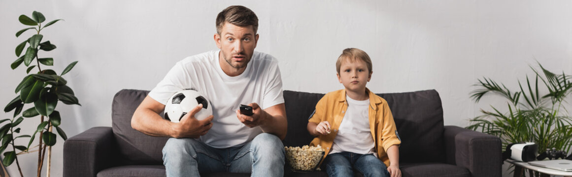 horizontal image of worried man holding soccer ball and tv remote controllers near son eating popcorn