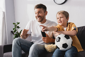 excited boy holding soccer ball and pointing with finger near cheerful father while watching tv