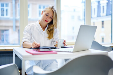 Pensive female entrepreneur making notes of working plans while having coffee break in office,...