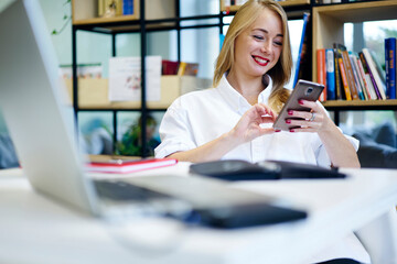 Cheerful business woman satisfied with successful project receiving congratulatory message on smartphone, young blonde student having fun in library watching videos in social networks using wifi
