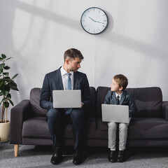 serious businessman and his son in formal wear sitting on sofa with laptops and looking at each...