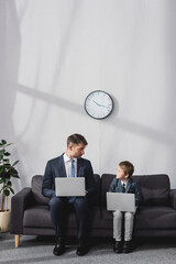 serious businessman and his son in formal wear sitting on sofa with laptops and looking at each...