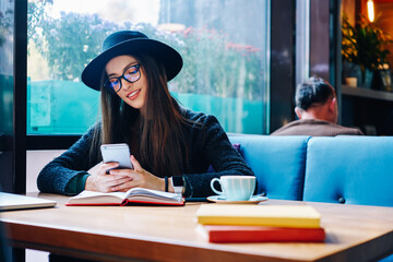 Attractive student writing email on smartphone and chatting online sitting in cafe.Beautiful female in stylish hat installing mobile application on cellular while preparing for exams in coffee shop