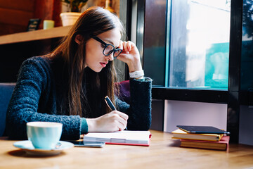 Talented brunette female student in eyewear learning in coffee shop writing report in notebook.Professional graphic designer pondering on creating sketch concentrated on work in cafe interior