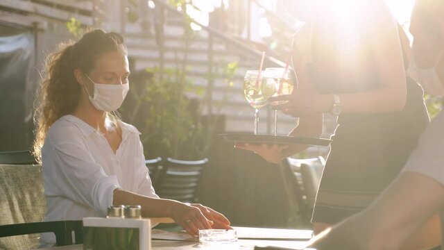 Waitress with protective medical mask serving guest with cocktails in outdoor restaurant. Opens new concept after quarantine. 4K