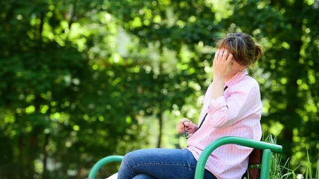 A woman sits in a Park and scratches her face from allergies.