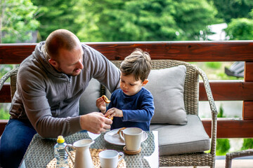 Young father is helping his son for breakfast. Dad and cute toddler boy having meal outdoor at the terrace. Smiling man with cheerful son having breakfast. Family day bonding.