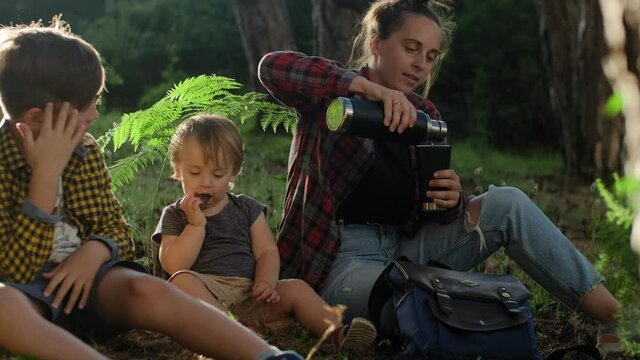 Mom With Two Sons Relaxing In The Forest And Drinking Hot Herbal Tea From A Thermos On A Sunny Day. Stay Cation 
