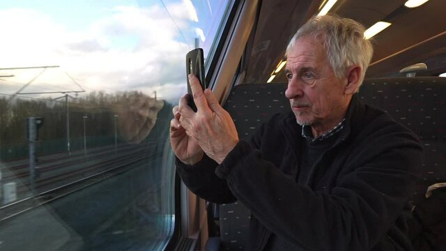 Man taking mobile photo through the train window. Mature caucasian man or senior citizen passenger holding smartphone and taking picture while sitting. Travelling or riding in public transport 