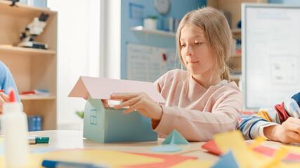Elementary School Arts & Crafts Class: Cute Little Blond Girl Has Fun with Her Classmates on the Handicraft Project, Uses Colorful Paper, Scissors and Glue to Build House