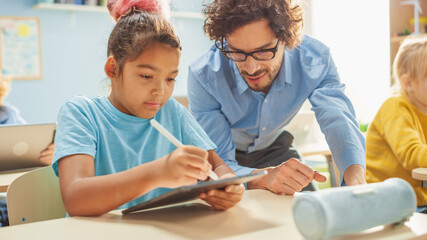 Elementary School Computer Science Class: Cute Girl Uses Digital Tablet Computer, Friendly Teacher Helps Her with the assignment. Children Getting Modern Education in STEM, Playing and Learning