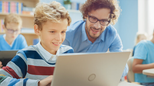 Elementary School Computer Science Class: Smart Boy Uses Laptop Computer, Friendly Teacher Helps Him by Explaining Lesson. Children Getting Modern Education in STEM, Playing and Learning
