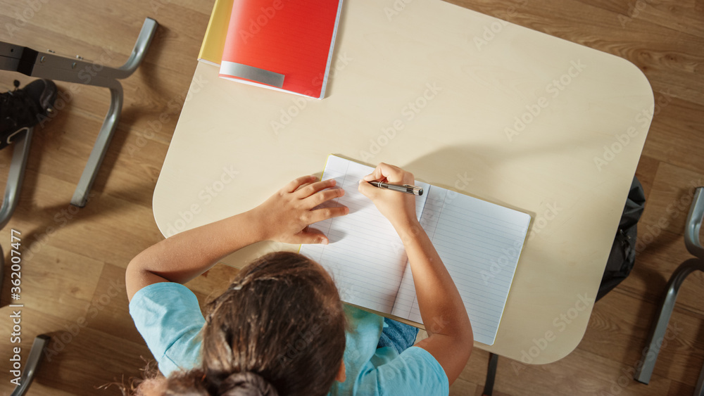 Poster top view shot of elementary school classroom: girl sitting at the school desk working on assignments