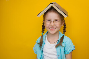 Back to school. Little beautiful girl in glasses with pigtails with an open book on her head smiles...
