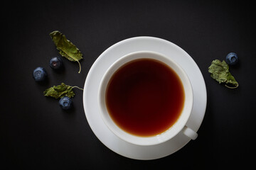 White cup with tea, blueberries and dried mint leaves on a dark background, view from above