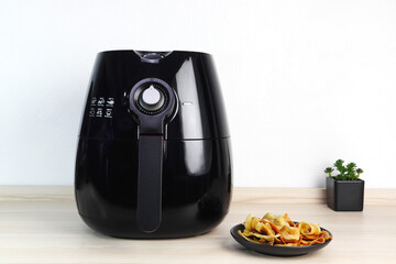 a black air fryer or oil free fryer appliance is on the wooden table in the kitchen with deep fried banan chips and small plant in the pot on white cement wall background during dinner in the party