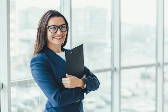 Confident young businesswoman standing with folder in hands, looking at the camera, ready to hard work day.