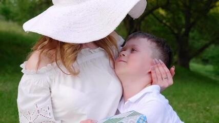 Mom in a white hat and son in a T-shirt are sitting on the grass in a park, hugging, kissing and smile on background of green trees. Woman holds in hand box with gift. 4K slow motion footage