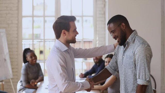 Proud caucasian manager boss handshaking congratulating praising male black african worker give positive feedback supporting promising reward promoting at team meeting, employee recognition concept