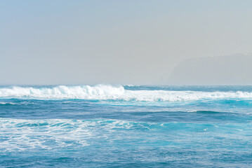 Blue ocean wave with white foam. Shoreline faded by the fog on background. Tenerife, Spain
