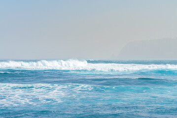 Naklejka na ściany i meble Blue ocean wave with white foam. Shoreline faded by the fog on background. Tenerife, Spain